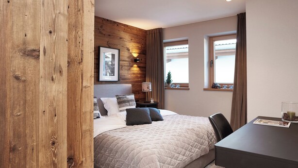 Quarto com cama, mesa de cabeceira, luminária e vista para o exterior pelas janelas.