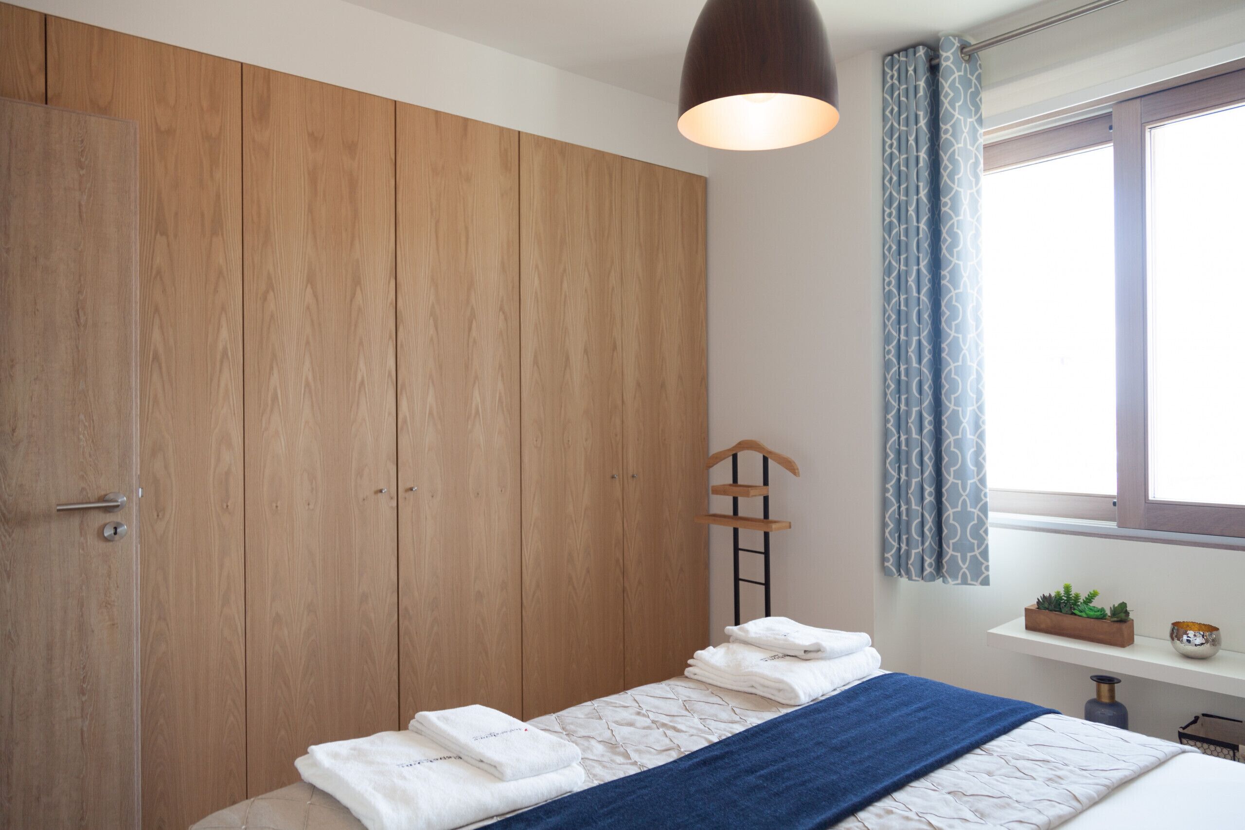 A bedroom with a wooden wardrobe, a bed with a blue blanket, a bedside table with a plant and a bowl, and a window with curtains.