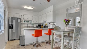 A modern kitchen with white cabinets, a stainless steel refrigerator, a coffee maker, and a dining area with a white table and orange chairs.