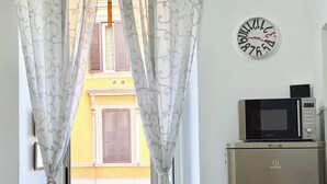 A view through a window with patterned curtains, a wall clock, and a microwave on a refrigerator. - Central Rome Apartment near Termini | Walk to Major Attractions | Roman Haven (Roma)