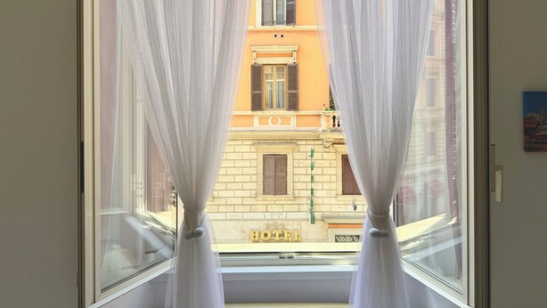 A window with sheer white curtains, a view of a building with a balcony and a sign that reads "HOTEL". - Central Rome Apartment near Termini | Walk to Major Attractions | Roman Haven (Roma)