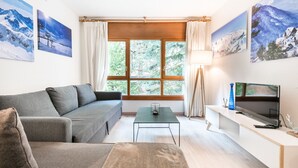Un salon moderne avec un canapé gris, une petite table basse, un téléviseur posé sur un meuble et une grande fenêtre avec des rideaux.