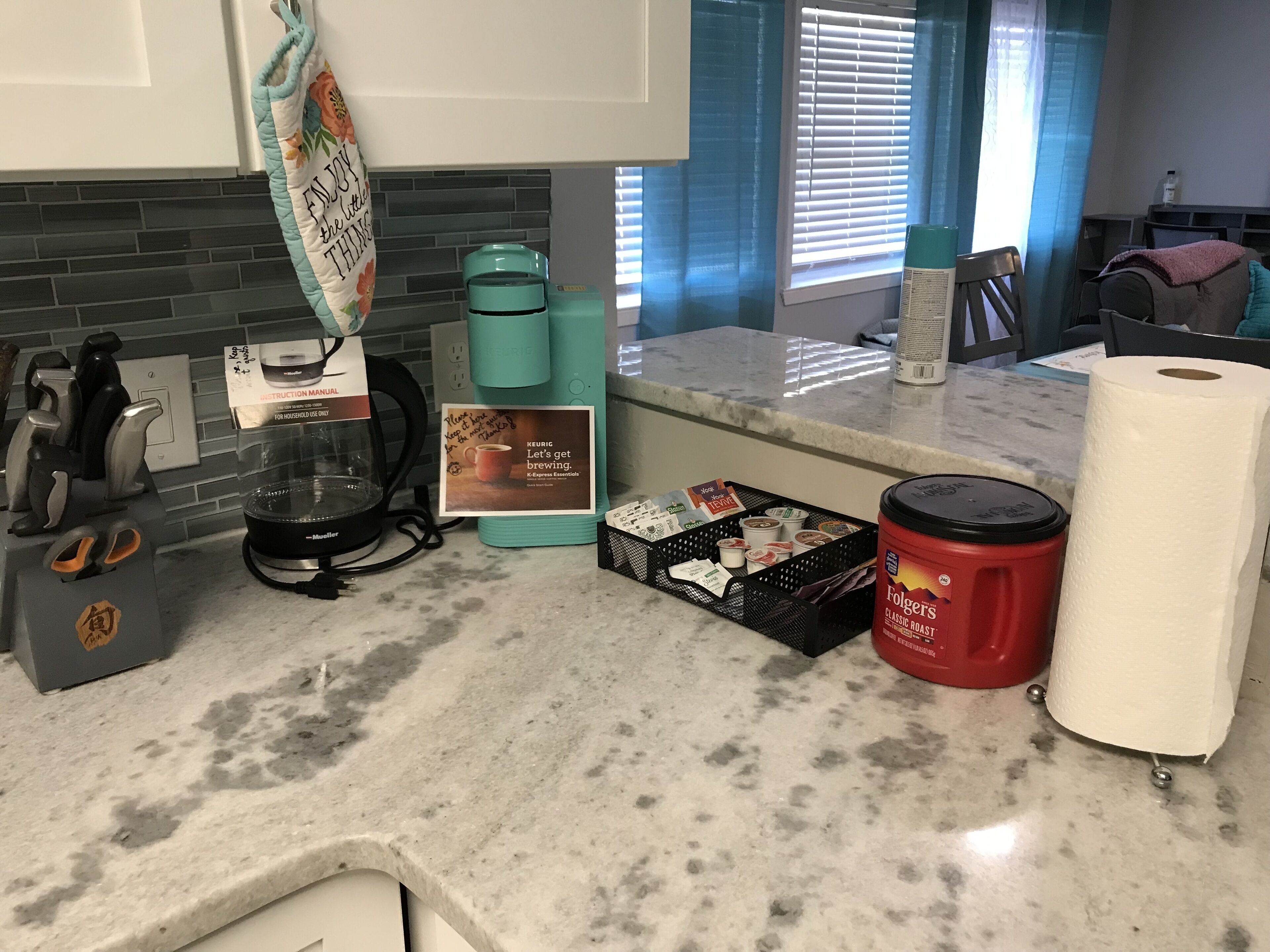 Una cocina con cafetera, un cesto de cápsulas de café, un recipiente rojo de Folgers y un rollo de toallas de papel.