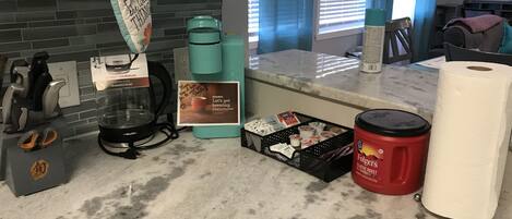 Una cocina con cafetera, un cesto de cápsulas de café, un recipiente rojo de Folgers y un rollo de toallas de papel.