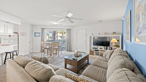 A living room with a sofa, coffee table, and TV. There is a dining area with chairs and a table. The room has white walls and a ceiling fan. - Shorehaven II 8B-2BR/2BA condo in Cherry Grove (North Myrtle Beach)