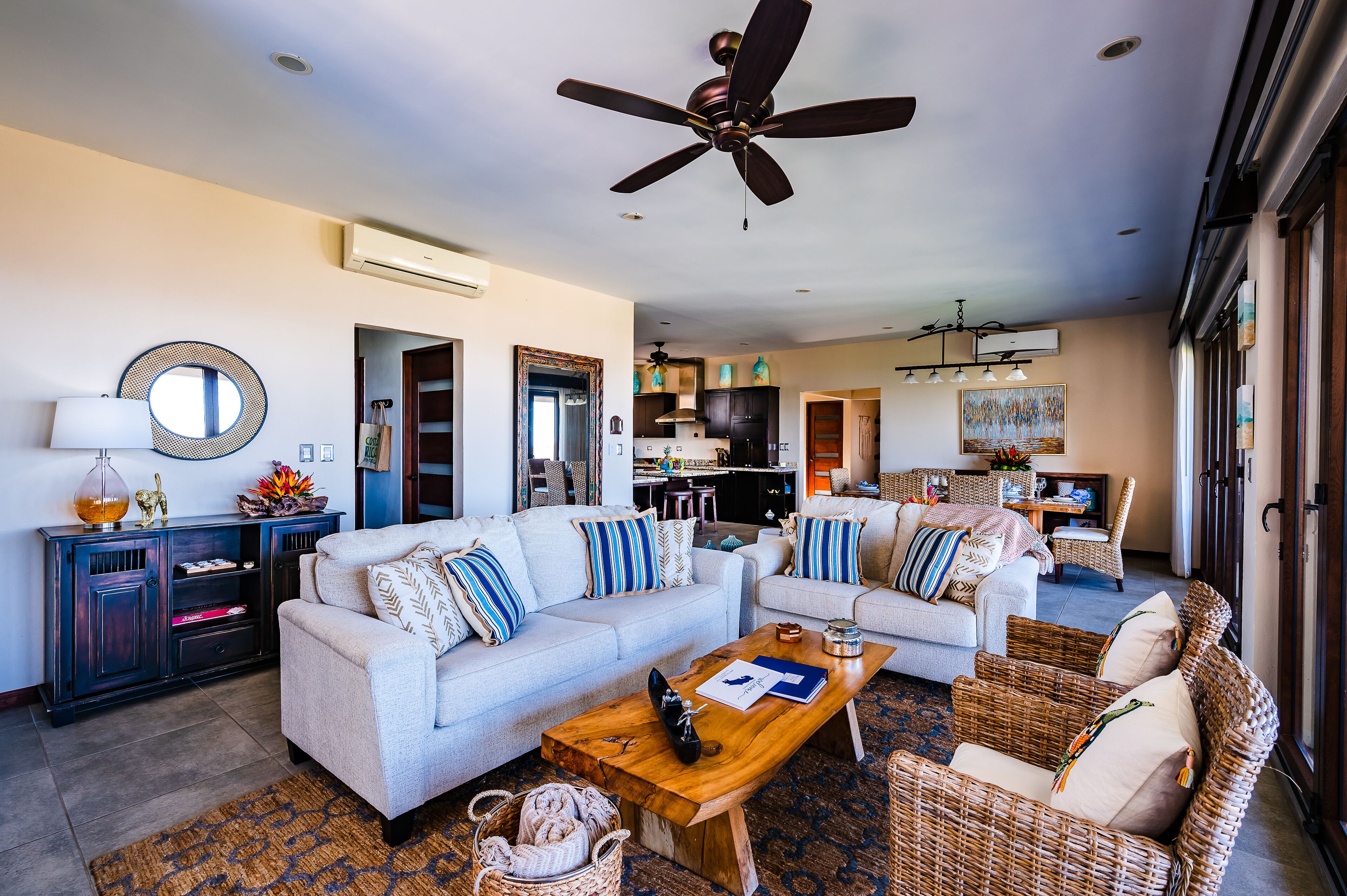 A spacious living room with a large sofa set, a wooden coffee table, and wicker chairs. There is a ceiling fan, a kitchen in the background, and a large window providing natural light.