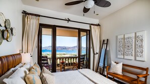Une chambre avec un grand lit, un banc en bois, un balcon donnant sur la mer et des œuvres d’art accrochées au mur.