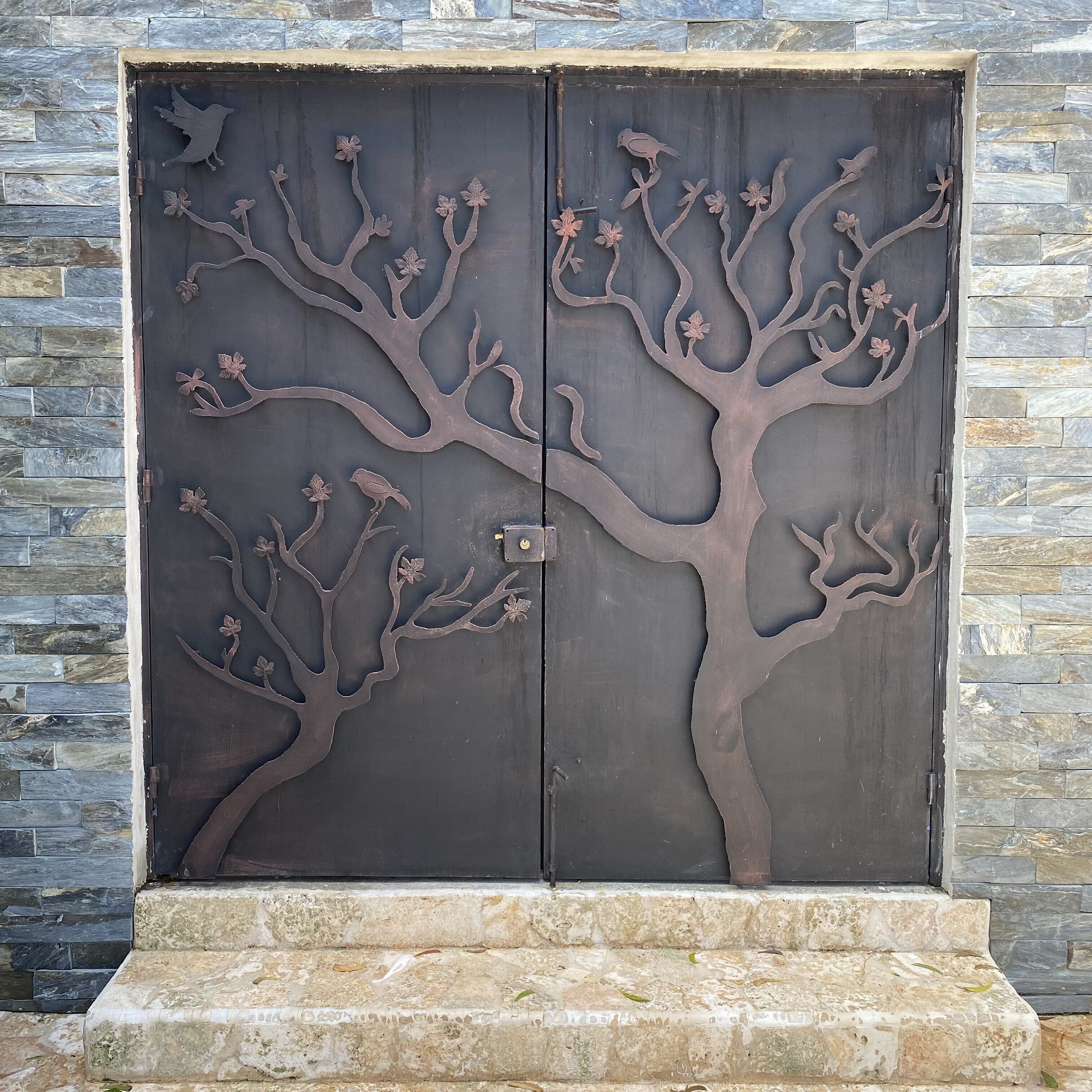 Eine Metalttür mit Baum- und Vogelmotiv vor einer Steinwand.