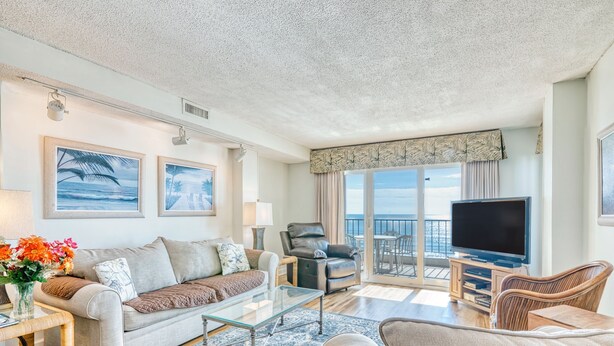 A spacious living room with a large sofa, a coffee table, and a television. There is a balcony with a view of the ocean.