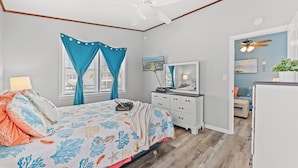 Une chambre à coucher comprenant un lit, un bureau, une chaise, un ventilateur de plafond et une fenêtre avec des rideaux.