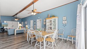 Un coin repas avec une table en bois et des chaises blanches, un ventilateur de plafond et une cuisine en arrière-plan.