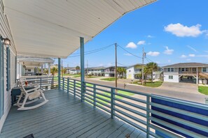 A covered porch with a rocking chair and a table, overlooking a residential street with houses and a clear sky.