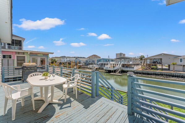 A wooden deck with a table and chairs, overlooking a calm water body and a row of houses.