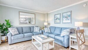 A living room with a light blue sofa, a white coffee table, and a wooden chair. There is a large mirror on the wall and two framed pictures.