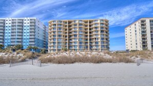 A beachfront hotel with multiple floors, a sandy beach in the foreground, and a clear blue sky.