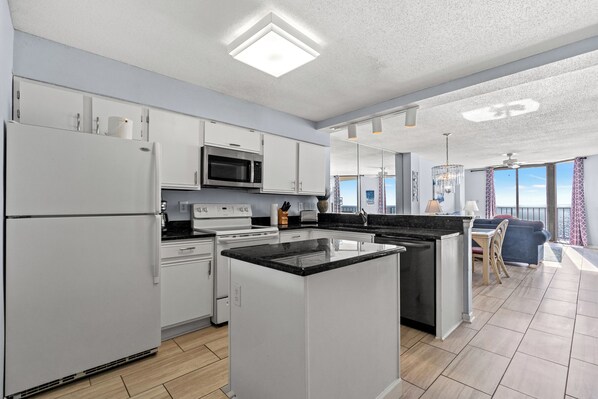 A modern kitchen with white cabinetry, a central island, and a dining area with a view. - Summit 9B - Ocean Front 3 Bed/3 Bath Condo - Accommodates 7 (North Myrtle Beach)