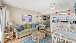 A living room with a sofa, a coffee table, and a dining table with chairs. There is a kitchen area with a refrigerator and a microwave.
