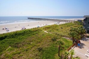A beach with a pier extending into the ocean, a grassy area in the foreground, and a building with a deck on the right.