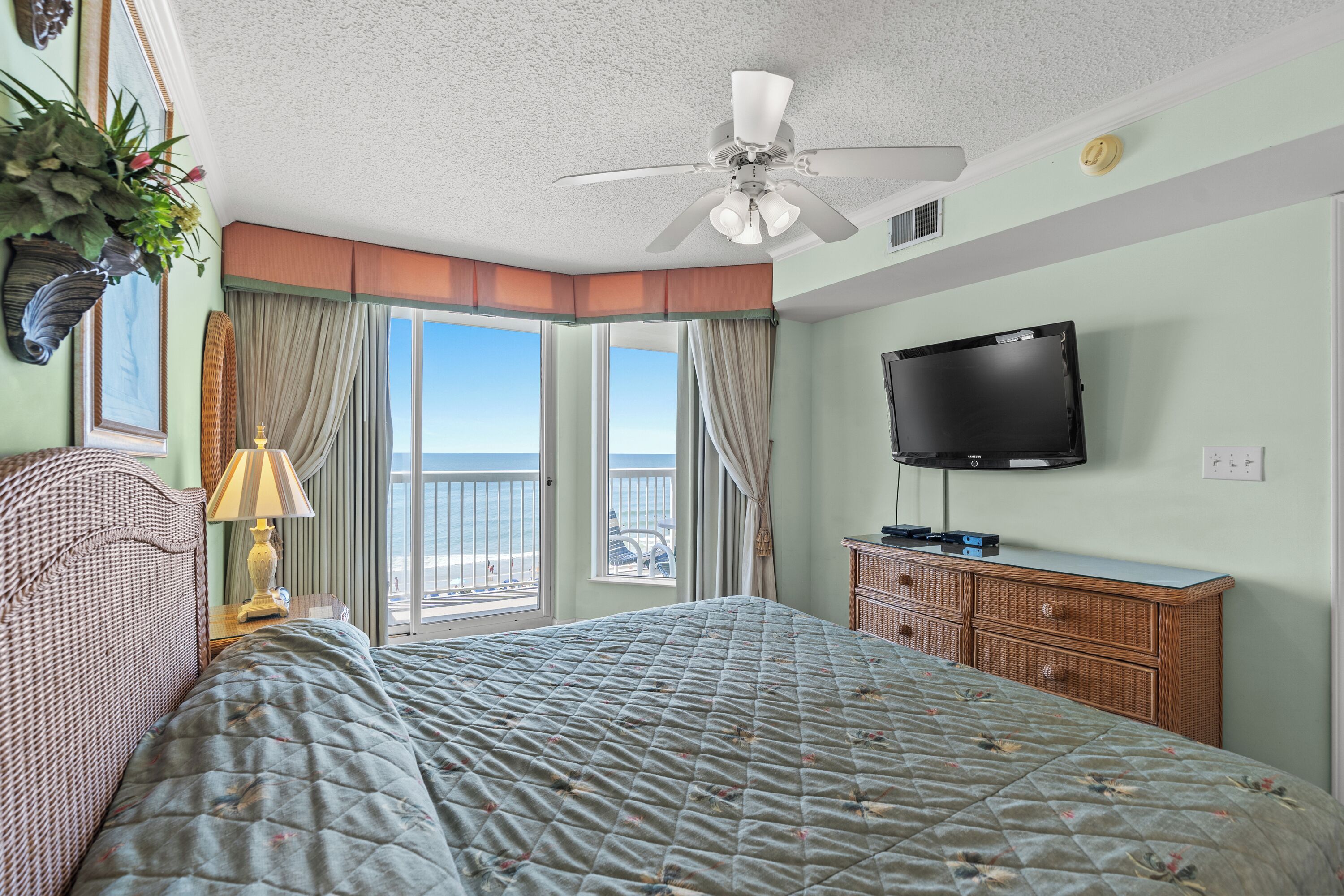 Ein Hotelzimmer mit Bett, Fernseher, Deckenventilator, einem Fenster mit Vorhängen und Blick auf den Strand.