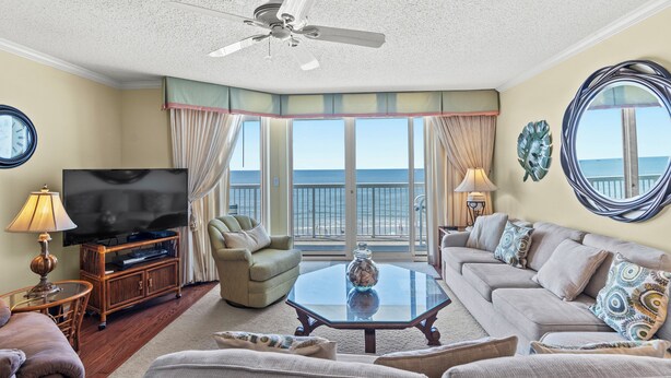 A spacious living room with a large flat-screen TV, comfortable seating, a ceiling fan, and a view of the ocean through large windows.