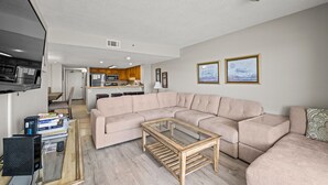 A modern living room with a beige sectional sofa, a glass coffee table, and a kitchen area in the background. - Sea Pointe 503 - Ocean Front Condo - Accommodates 7 in 3 Bedrooms & 2 Baths (North Myrtle Beach)