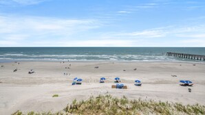 A beach with several blue umbrellas, a pier extending into the water, and a sandy shore. - Sea Pointe 503 - Ocean Front Condo - Accommodates 7 in 3 Bedrooms & 2 Baths (North Myrtle Beach)