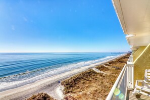 A beach view from a balcony with a wooden railing, a chair, and a clear sky.