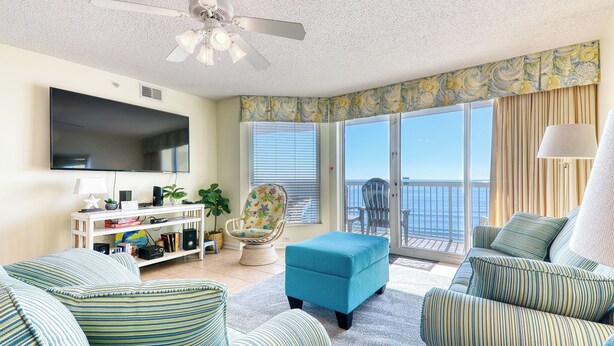 A living room with a flat-screen TV, a ceiling fan, a sofa, an ottoman, a chair, a TV stand, and a view of the ocean through a sliding glass door.