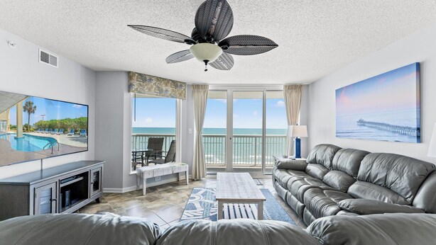 A living room with a ceiling fan, a television, a sofa, a coffee table, and a view of the ocean through large windows.