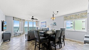 A spacious living area with a dining table, chairs, a ceiling fan, and large windows offering a view of the ocean.