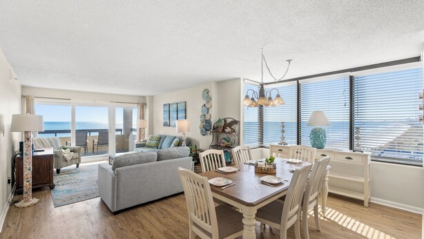 A spacious living area with a dining table, chairs, and a sofa. There is a large window with blinds, a chandelier, and artwork on the walls. - Sea Pointe 908 -Oceanfront 3BR/2BA Condo (North Myrtle Beach)