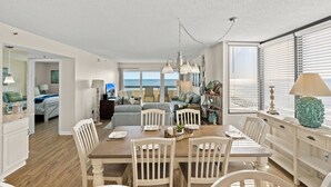 A spacious living area with a dining table set for four, a large window with blinds, and a view of the ocean. - Sea Pointe 908 -Oceanfront 3BR/2BA Condo (North Myrtle Beach)