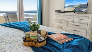 A hotel room with a bed, a TV displaying a beach view, a dining area with chairs, and a view of the ocean. - Sea Pointe 908 -Oceanfront 3BR/2BA Condo (North Myrtle Beach)