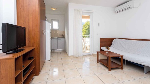 Un espacio habitable compacto con un televisor sobre un soporte de madera, un sofá, una mesita y una cocina visible a través de una puerta abierta.