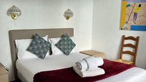 A neatly made bed with white linens, a red throw blanket, and patterned pillows. There is a wooden nightstand with a lamp, a wooden chair, and a wall art depicting a person driving.