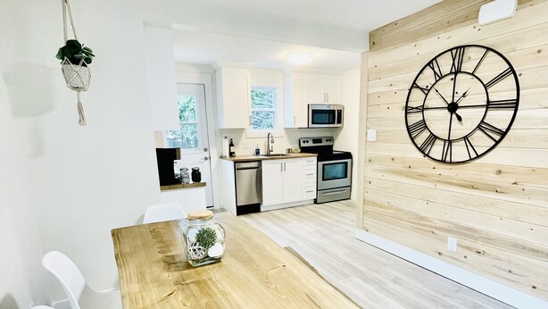 A modern kitchen with white cabinets, a wooden dining table with black legs, and a large wall clock. - The Nest - A Shore Paradise (Pointe-du-Chêne)