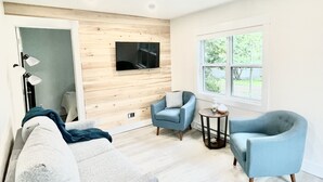 A modern living room with a grey sofa, two blue armchairs, a wooden wall panel, a flat-screen TV, and a large window with a view of greenery. - The Nest - A Shore Paradise (Pointe-du-Chêne)