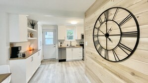 A modern kitchen with white cabinets, a wooden wall clock, and a view into the dining area. - The Nest - A Shore Paradise (Pointe-du-Chêne)