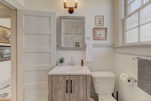 A bathroom with a white sink, a toilet, and a mirror above it. There are two framed pictures on the wall and a window with a view of trees.