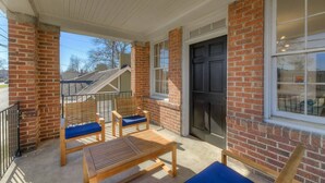 A covered porch with brick walls, a black door, and two wooden chairs with blue cushions. - The Jewel of Bull Street Upstairs - 1 King Bedroom (Columbia)