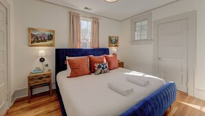 A bedroom with a large bed, a headboard, a nightstand, a window with curtains, and a door. - The Jewel of Bull Street Upstairs - 1 King Bedroom (Columbia)