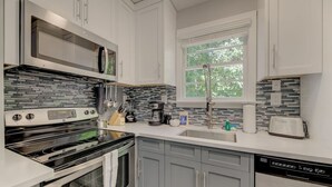 A modern kitchen with white cabinets, a stainless steel range, a dishwasher, and a window with a view of trees.