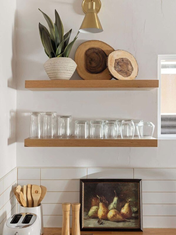 A kitchen with wooden shelves holding glass jars, a potted plant, and a wooden log. Below, there is a toaster, wooden utensils, and a framed painting of pears.