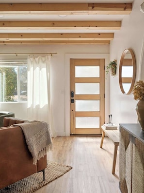 Una sala de estar acogedora con techo de madera, un ventanal con cortinas, un sofá marrón, una mesita con un florero y una puerta de madera con paneles de vidrio esmerilado.