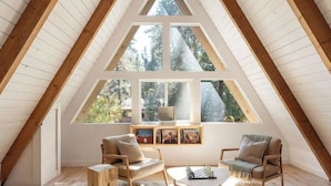 Um quarto de sótão aconchegante com teto inclinado, vigas de madeira e janelas amplas com vista para árvores. Há duas cadeiras, uma mesa pequena e uma estante com quadros.