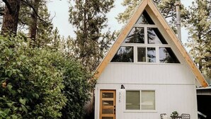 Une petite cabane blanche en forme de «A» avec une porte et des fenêtres en bois, entourée d’une végétation luxuriante.