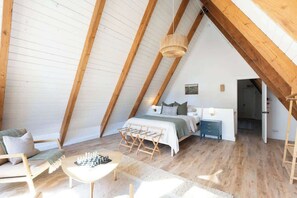 A cozy attic bedroom with a sloped ceiling, wooden beams, a bed with a green blanket, a chessboard, and a small table.
