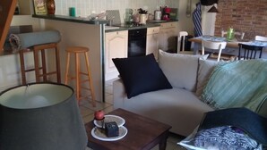 A cozy living room with a sofa, coffee table, and a lamp. Adjacent is a kitchen with white cabinets and a dining area with a table and chairs.