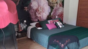 A bedroom with a bed, a desk, and a chair. There is a floral tapestry on the wall and a painting on the floor.