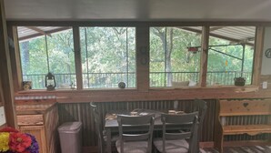 A rustic wooden room with a table and chairs, large windows with a view of greenery, and a small shelf with decorative items.